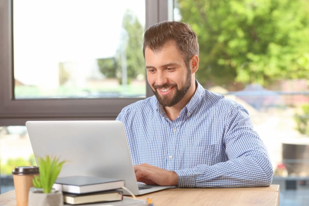 man sitting at a desk indoors at his computer smiling. he is wearing a checkered shirt and there is a window behind him. Through the window you can see a tree